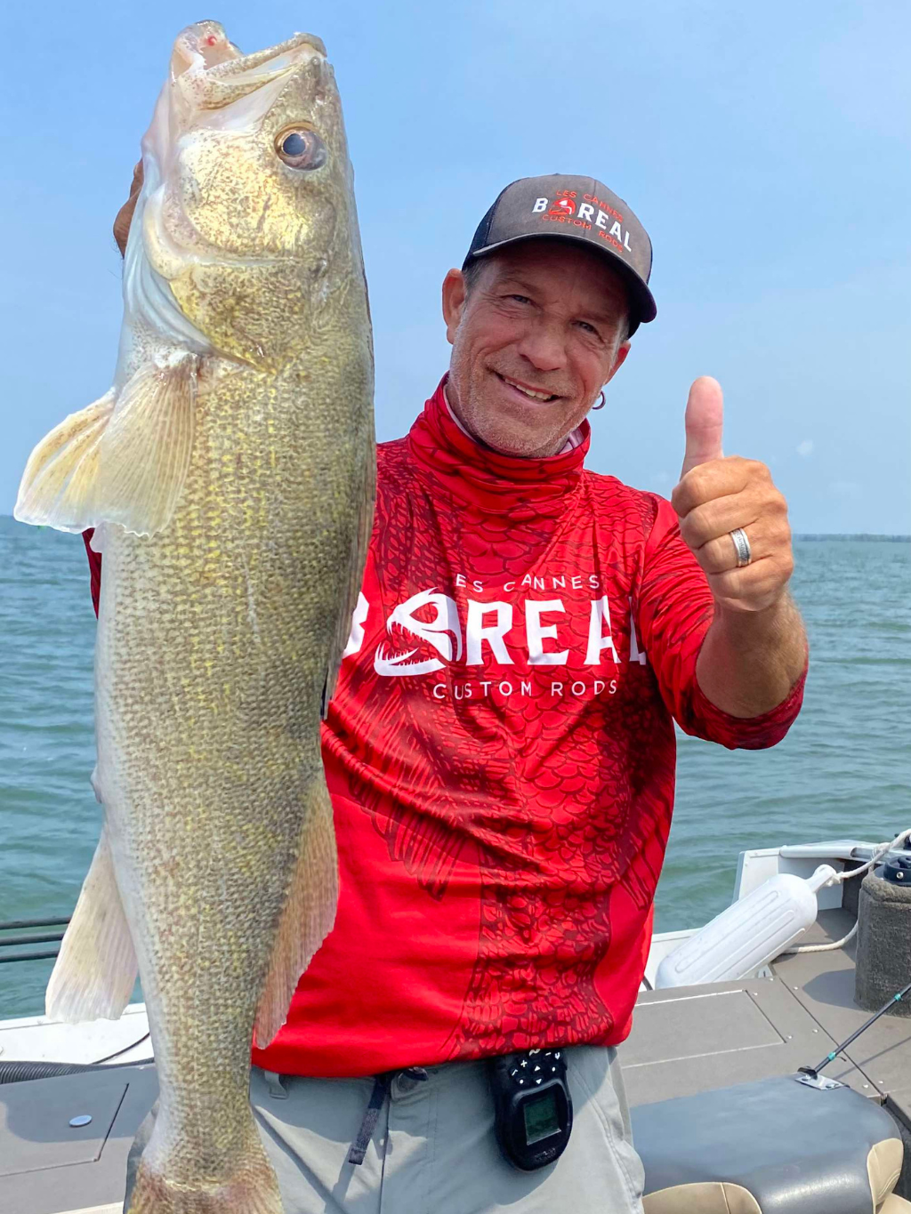 Pêcheur souriant sur un bateau tenant un doré trophée, vêtu d’un chandail rouge Les Cannes Boréal Custom Rods.